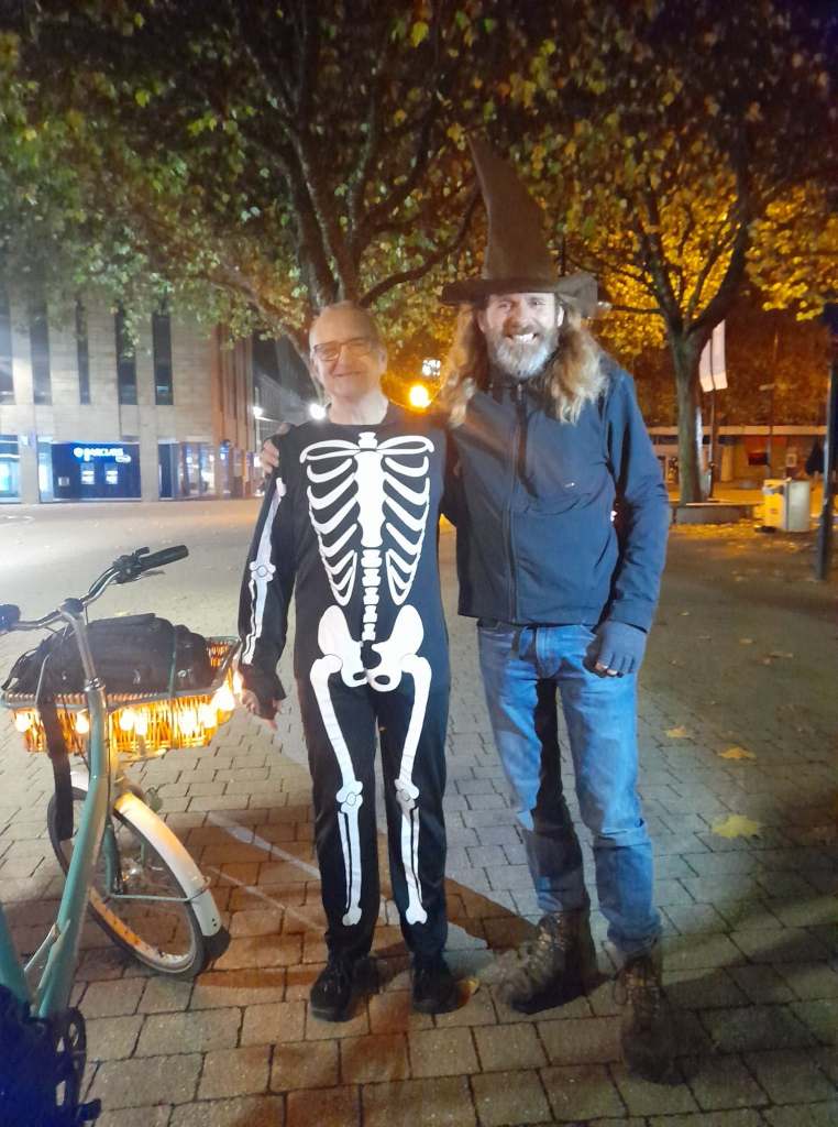 A man with a skeleton suit and one with wearing a wizard hat, standing next to an Elephant Bike with pumpkin lights around the front basket. (Thanks to Sadie Tann)
