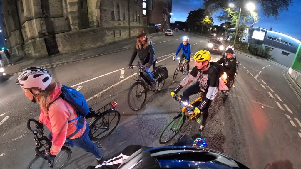 Halloween themed cyclists waiting to set off from traffic lights. There is a car waiting behind them.