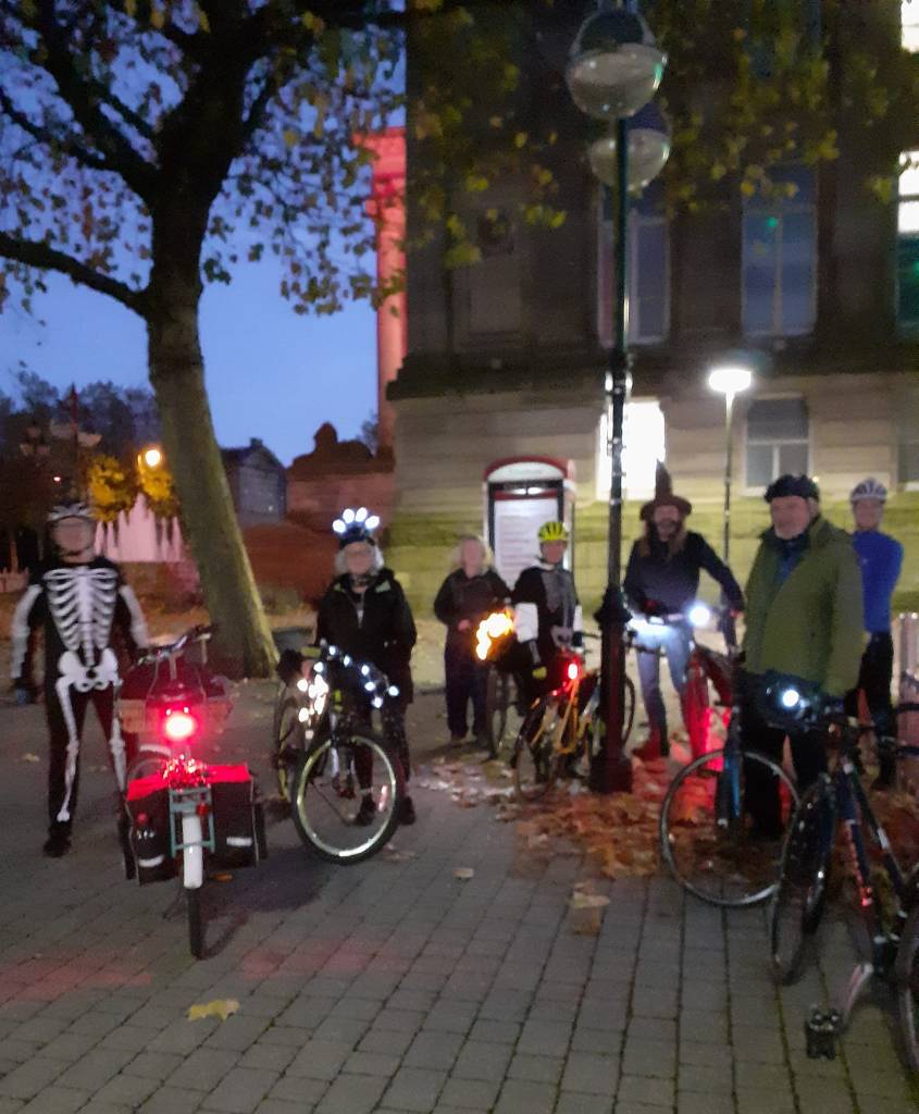 A group of people with bicycles, many dressed in Halloween costumes and with spooky lights on their bicycles. (Thanks to June Leece)