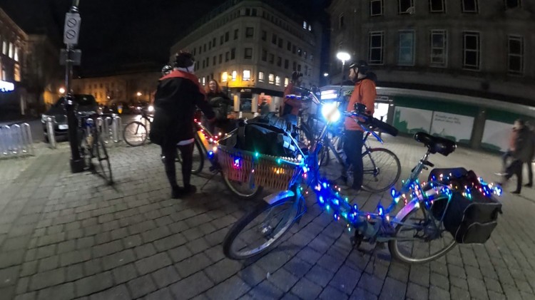 A collection of bicycles are parked on Victoria Square in Bolton at night and the riders are chatting. A small cargo bicycle in the foreground is decorated with coloured lights.
